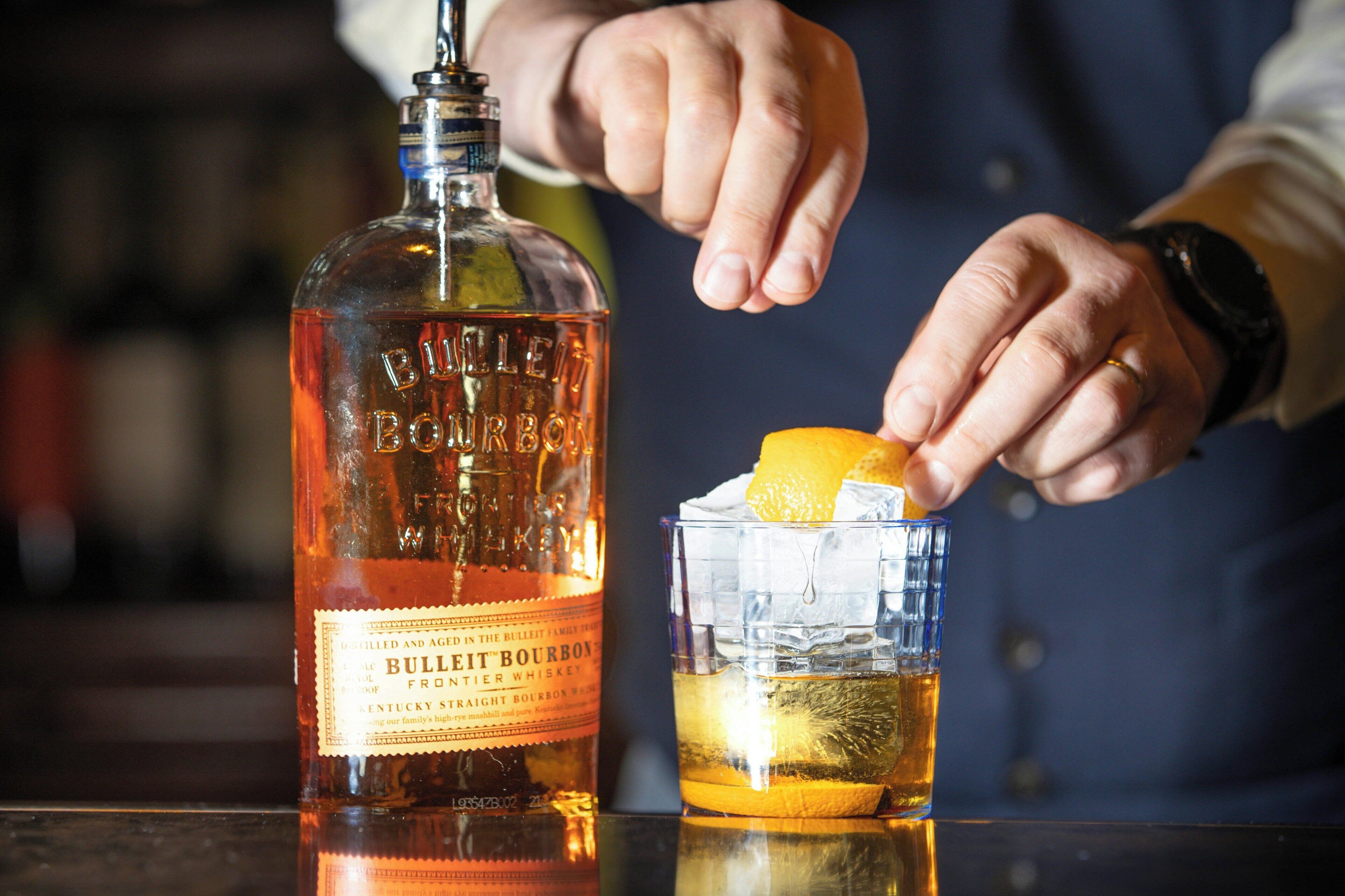 A person pouring a drink from a bottle labeled "BULLITE BOURBON" into a glass with ice and an orange slice.