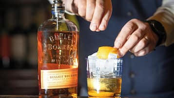 A person pouring a drink from a bottle labeled "BULLITE BOURBON" into a glass with ice and an orange slice.