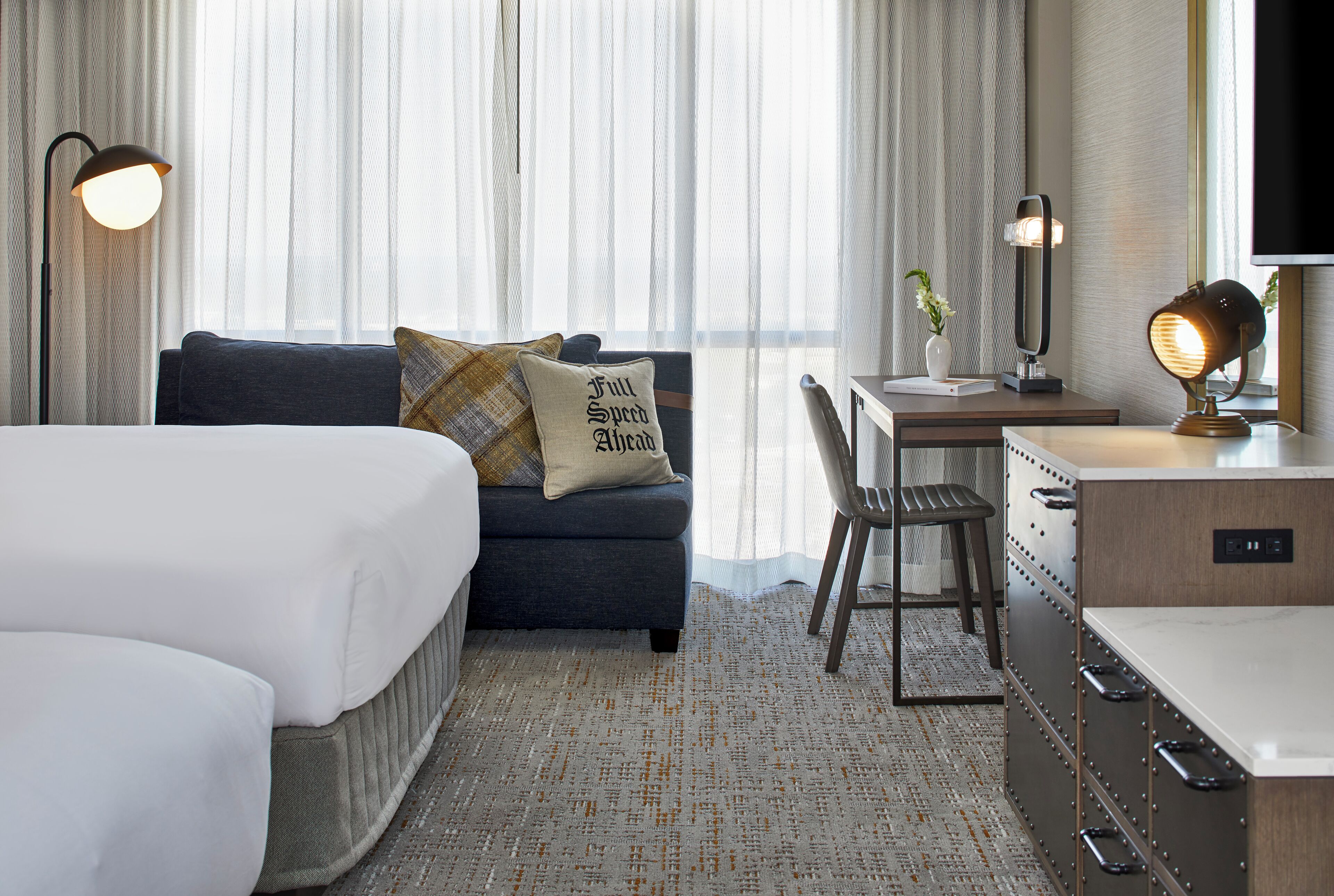 A hotel room with a bed, a sofa with pillows, a desk with a chair, a mirror, and a lamp.