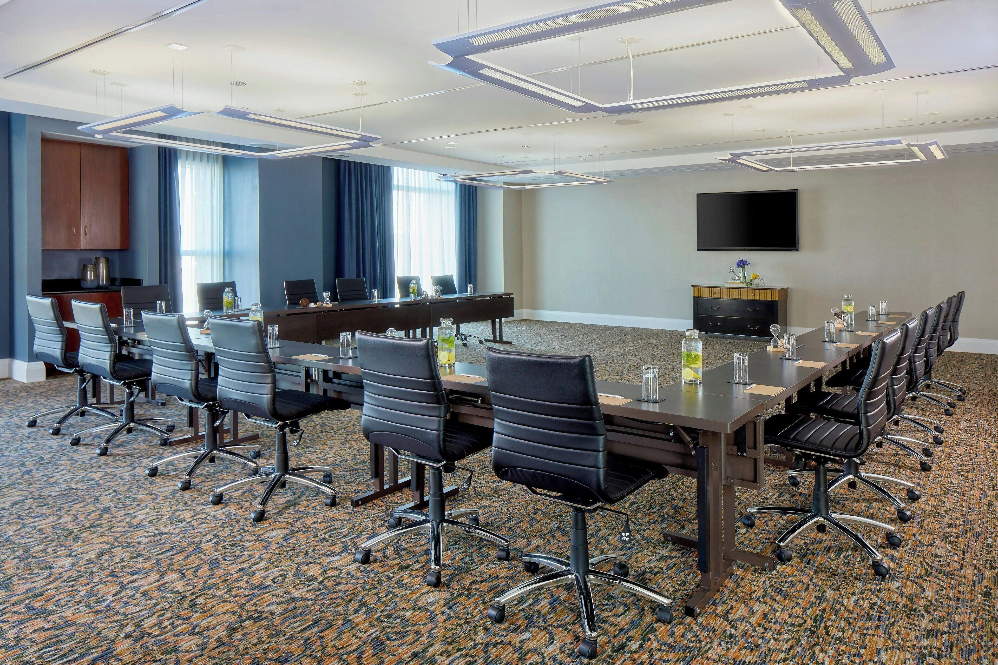 A conference room with a long table, multiple chairs, a flat-screen TV, and a large window with curtains.