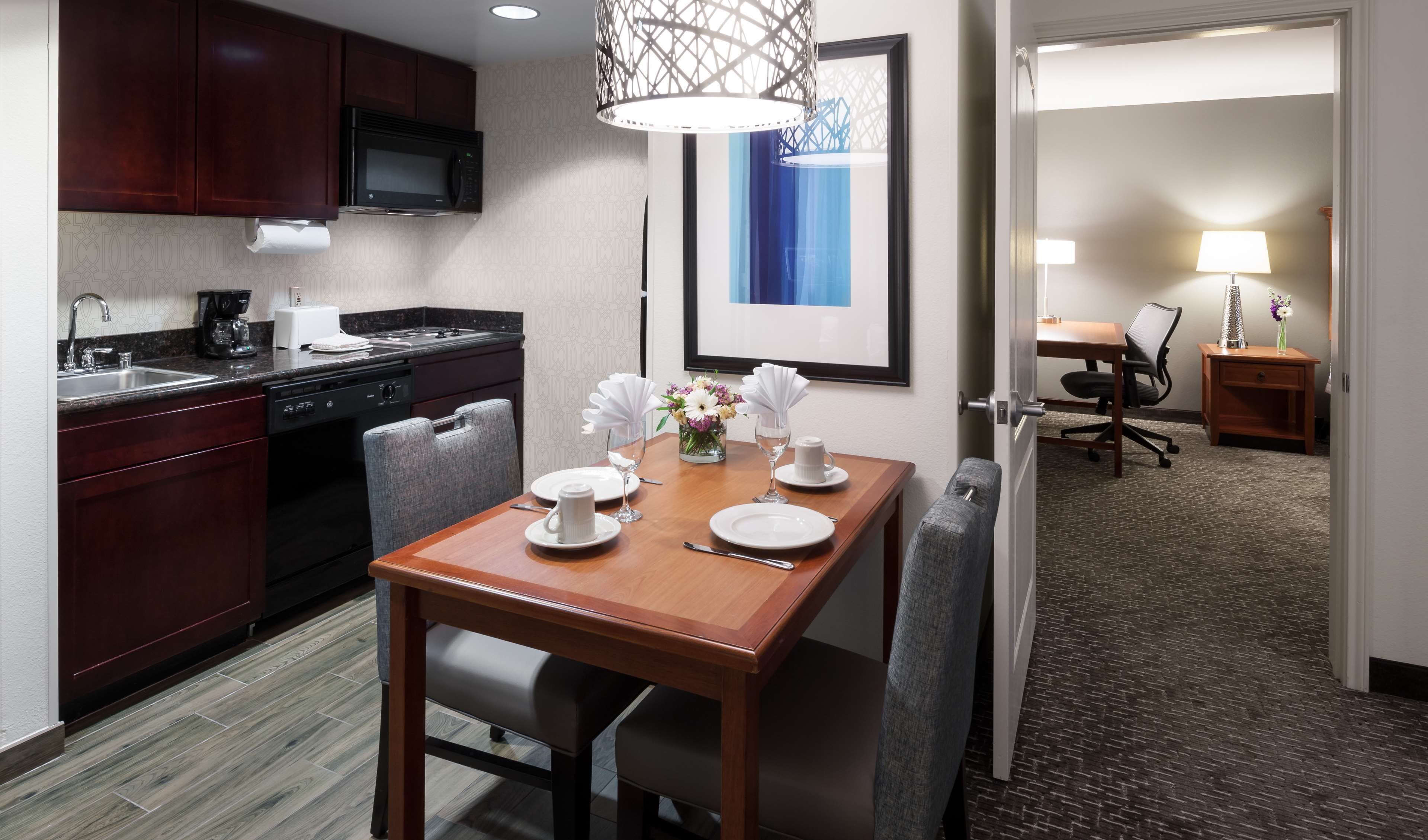A hotel room with a dining area featuring a wooden table set for two, a microwave, a coffee maker, and a sink.
