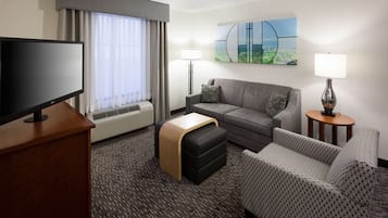 Ett hotellrum med en platt-TV, en soffa, en mindre soffa, ett litet bord, en lampa, en värmare och ett fönster med gardiner.