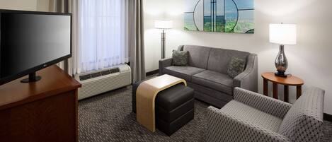 A hotel room with a flat-screen TV, a sofa, a loveseat, a small table, a lamp, a heater, and a window with curtains.
