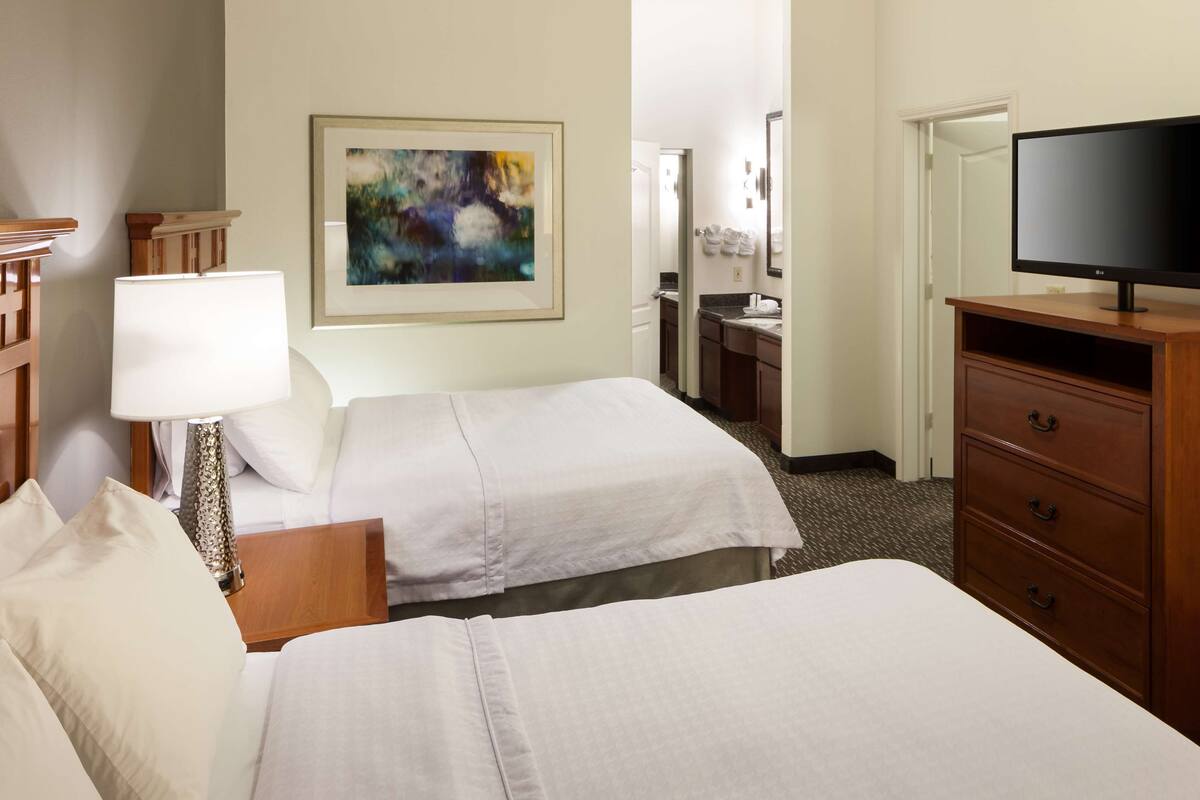 Une chambre d’hôtel avec deux lits, un bureau avec une lampe, une commode, un téléviseur et un tableau au mur.