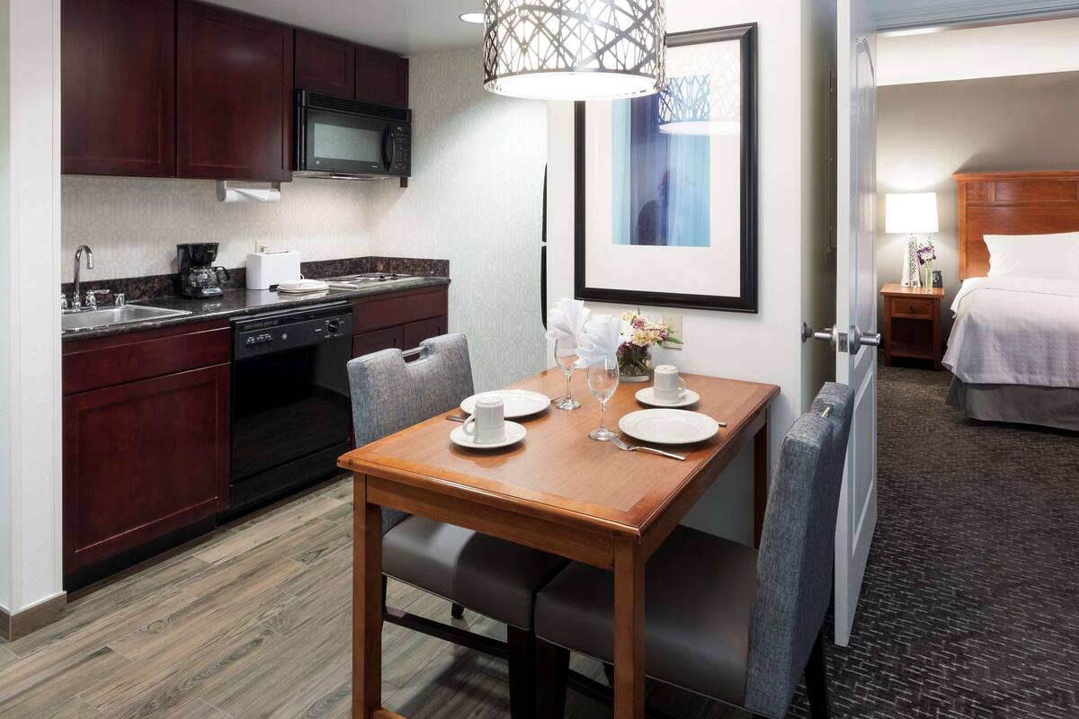 Une chambre d’hôtel équipée d’une kitchenette, d’une table à manger dressée pour deux personnes, d’un lit et d’une salle de bain visible à travers une porte ouverte.
