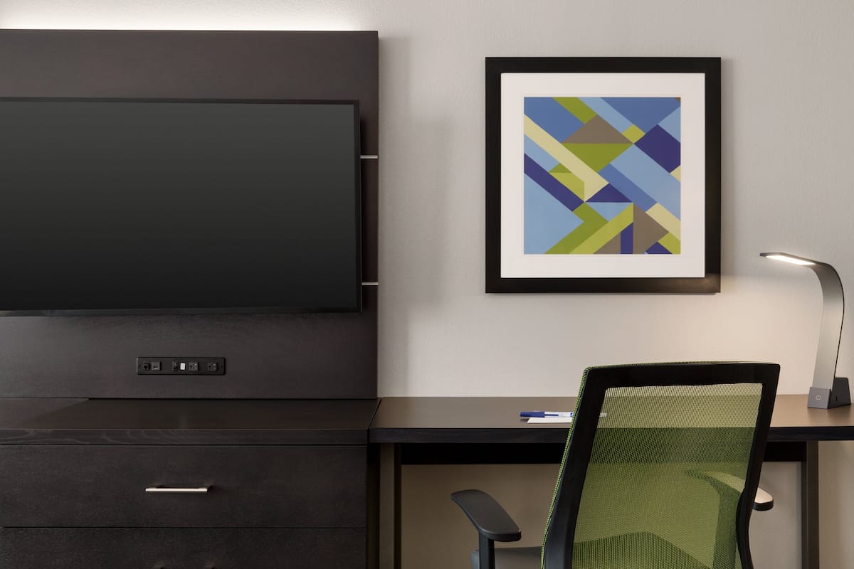 Un espacio de oficina moderno con una televisión de pantalla grande, una obra de arte abstracta enmarco, un escritorio con una silla y una lámpara de escritorio.