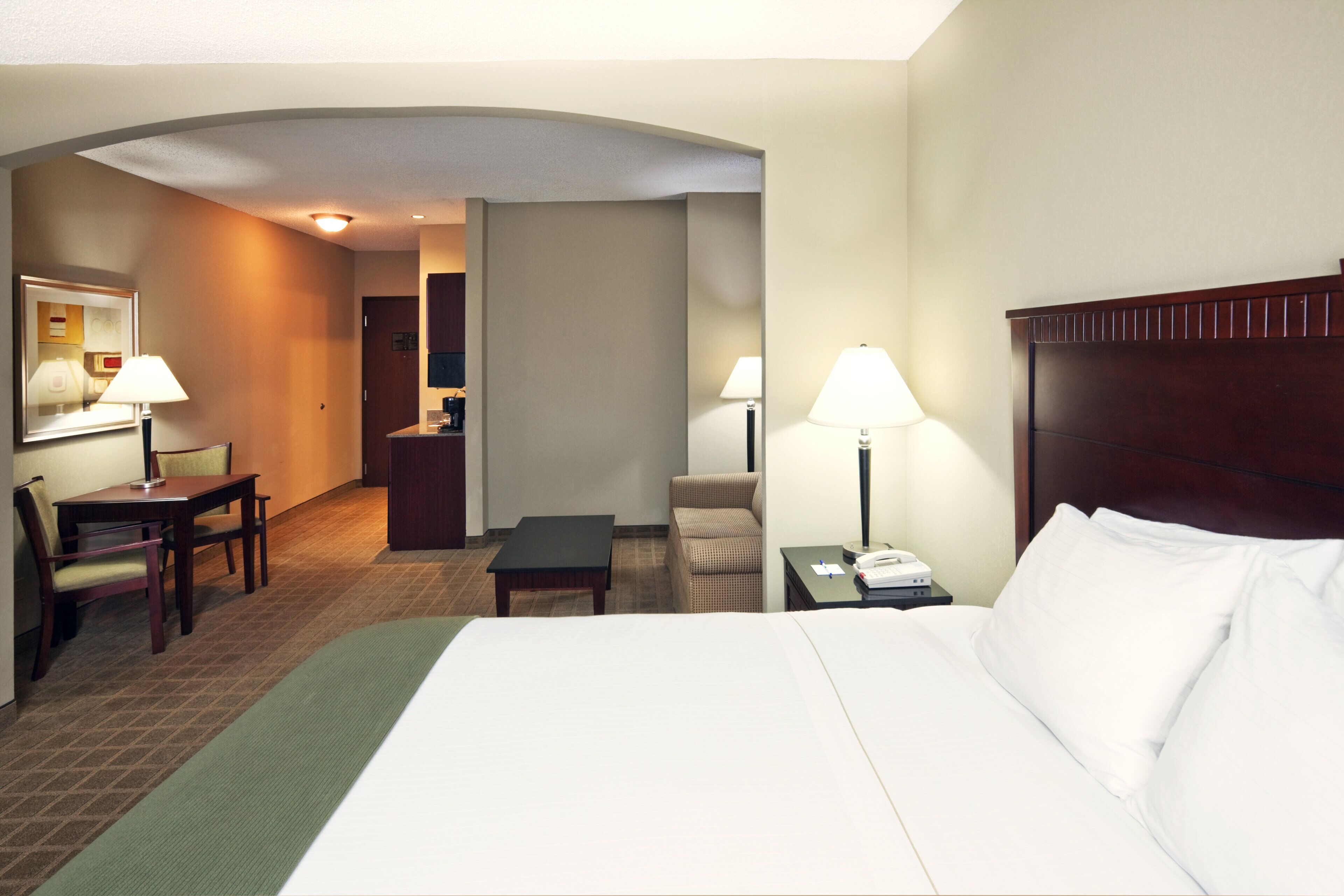 Habitación de hotel con una cama, dos mesitas de noche con lámparas, un escritorio con una silla y una pequeña mesa con otra silla.