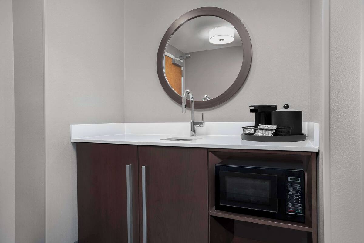 A modern bathroom with a white sink, a round mirror, a coffee maker, and a microwave.