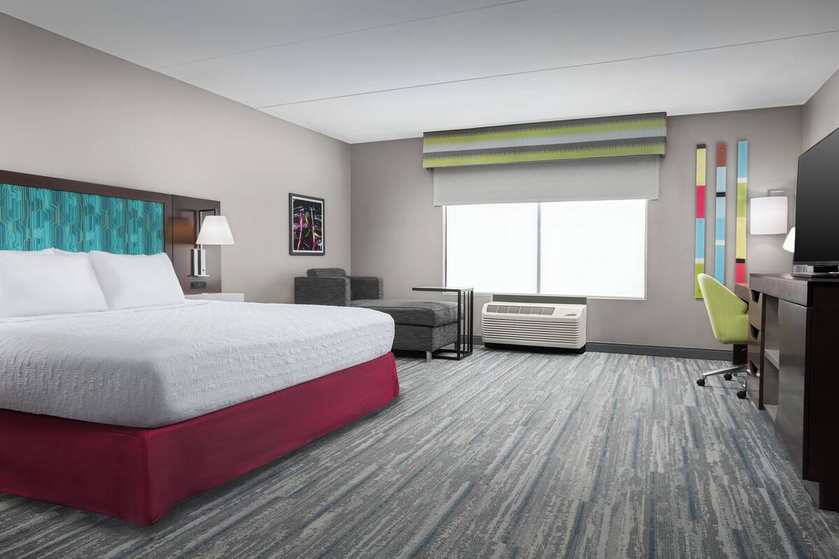 A hotel room with a bed, a desk, a chair, an air conditioner, and a window with blinds.
