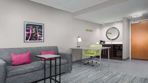 Ein modernes Hotelzimmer mit grauem Sofa, einem kleinen Tisch, einem grünen Bürostuhl, einem Schreibtisch mit Lampe, einer Mikrowelle und einer Holztür.