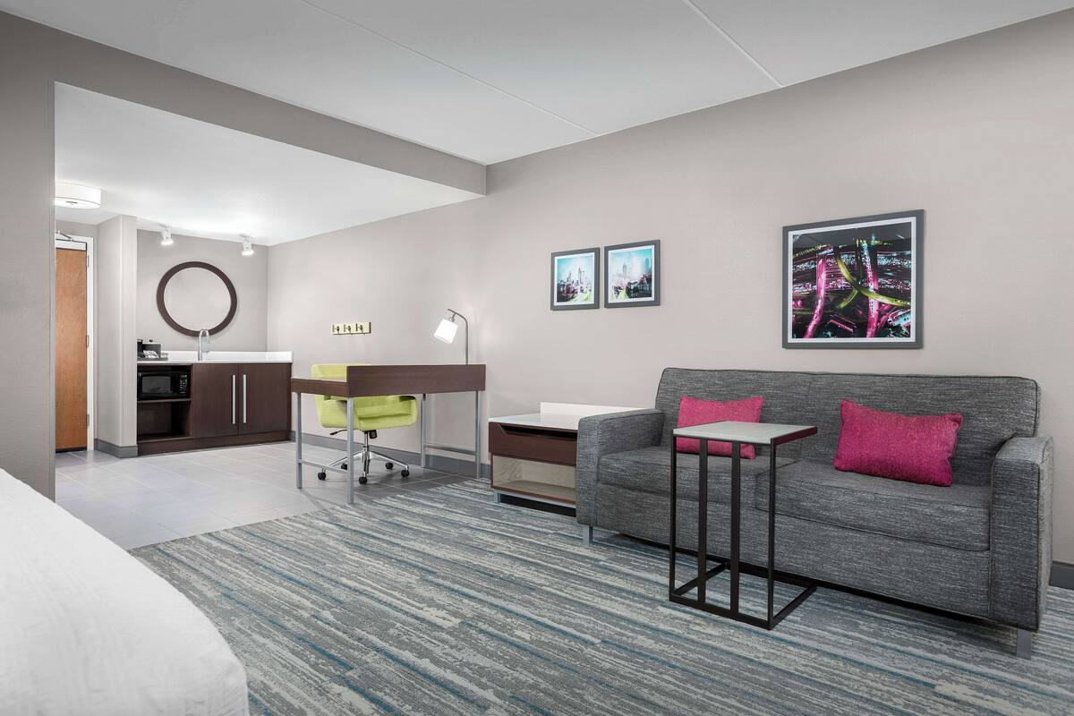 A modern hotel room with a grey sofa, a small table, a desk with a lamp, a microwave, and a ring light. There are two framed pictures on the wall and a carpeted floor.