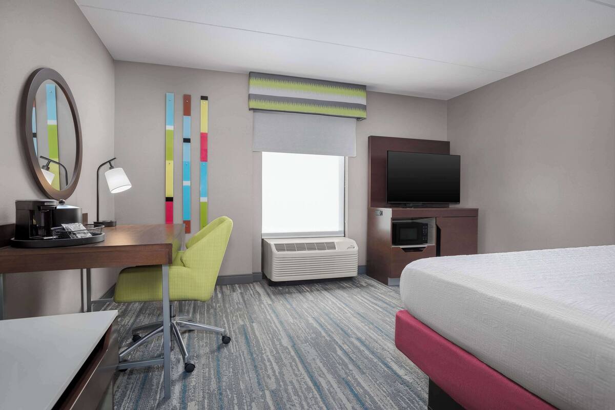 A hotel room with a bed, desk, chair, TV, air conditioning unit, and a window.