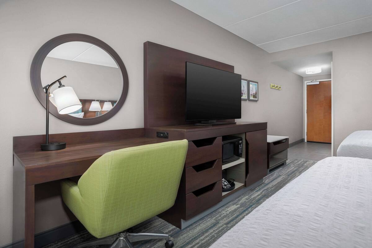A hotel room with a bed, a desk with a chair, a television, a mirror, and a door.
