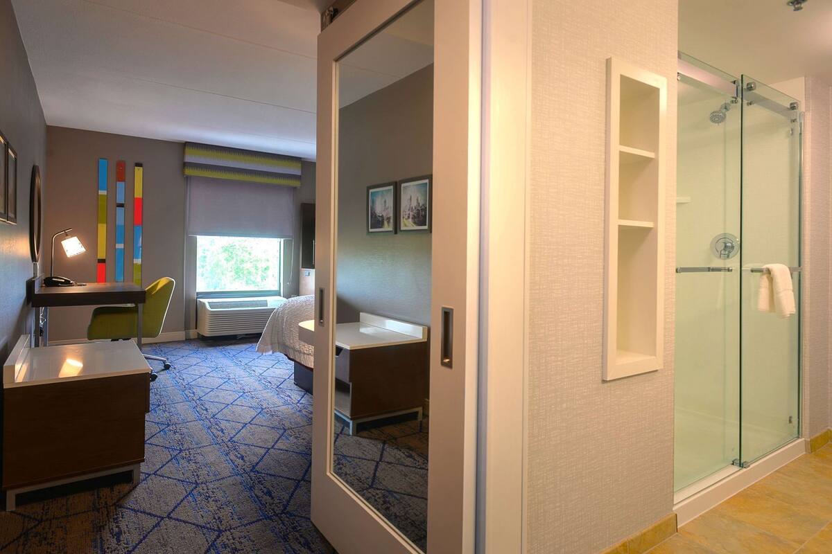 A hotel room with a desk, chair, bed, and a glass-enclosed shower.