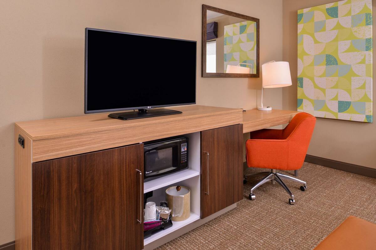 A hotel room with a wooden desk, a flat-screen TV, a microwave, a lamp, and an orange chair.