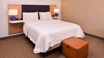 A hotel room with a large bed, two bedside tables with lamps, a brown ottoman, and a wooden headboard.