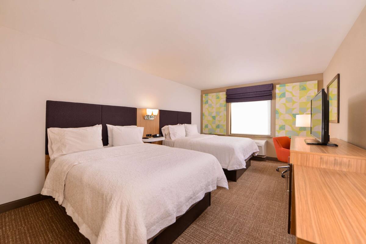 A hotel room with two beds, a desk, a chair, and a TV.