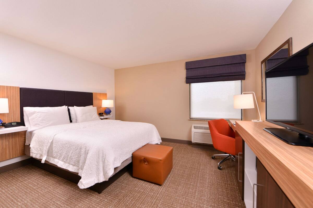 A hotel room with a bed, bedside tables, a desk, a chair, a window with blinds, and a mirror.