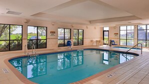 Una piscina interior con vistas despejadas a través de amplios ventanales, una silla de salvavidas y una zona junto a la piscina con una mesa y sillas.