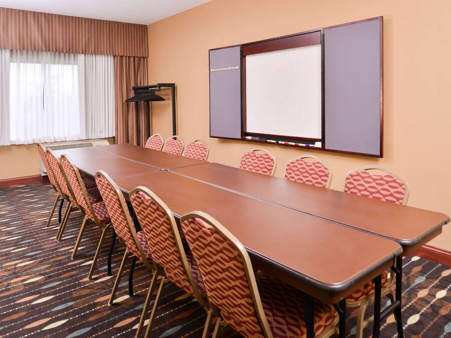 A conference room with a long table, six patterned chairs, a projector screen, and a carpeted floor.