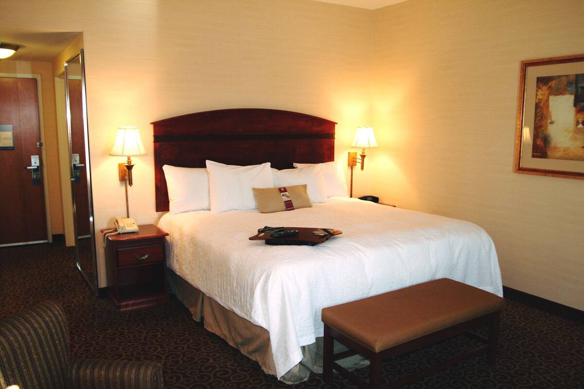 A hotel room with a large bed, two bedside lamps, a bedside telephone, a bench, and a painting on the wall.