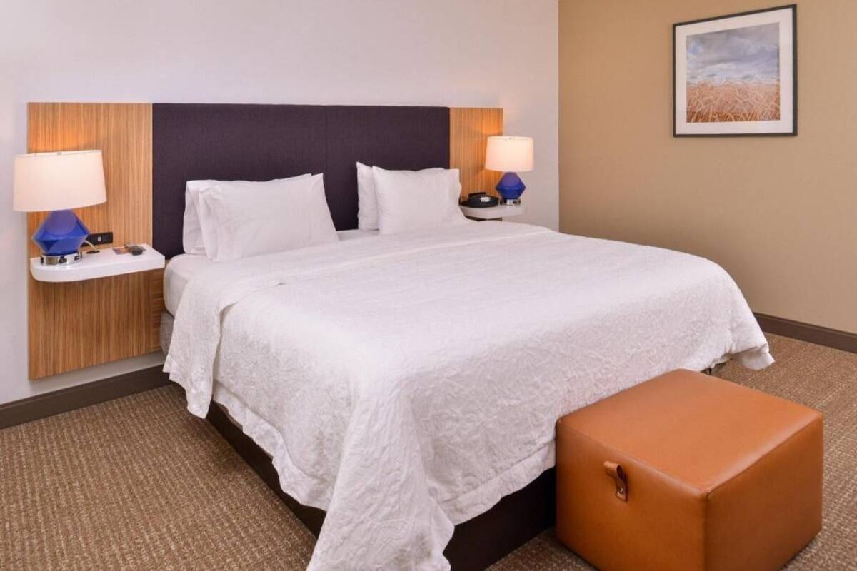 A hotel room with a bed, two bedside tables with lamps, a nightstand, a brown ottoman, and a framed picture on the wall.