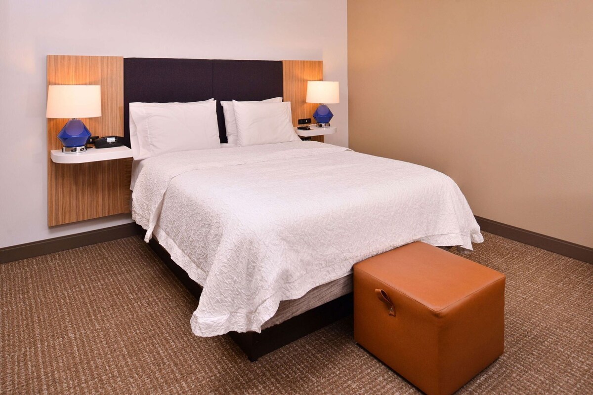A hotel room with a bed, two bedside tables with lamps, a brown ottoman, and a textured carpet.