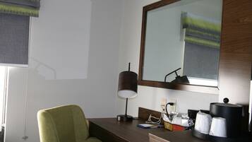 Une chambre d’hôtel avec un fauteuil vert, un bureau avec une lampe, une machine à café et un miroir qui reflète une fenêtre avec des stores.