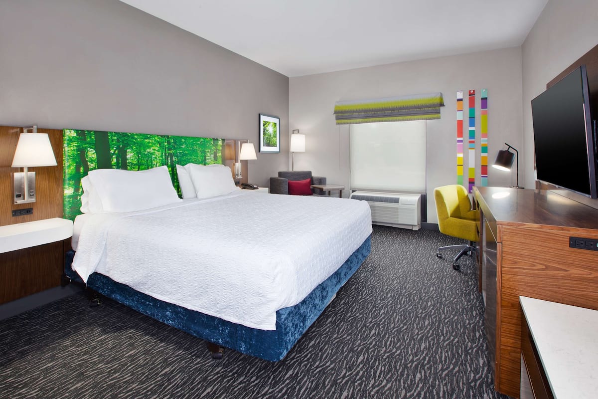 A hotel room with a large bed, a desk, a chair, a TV, and a window with a view of trees.