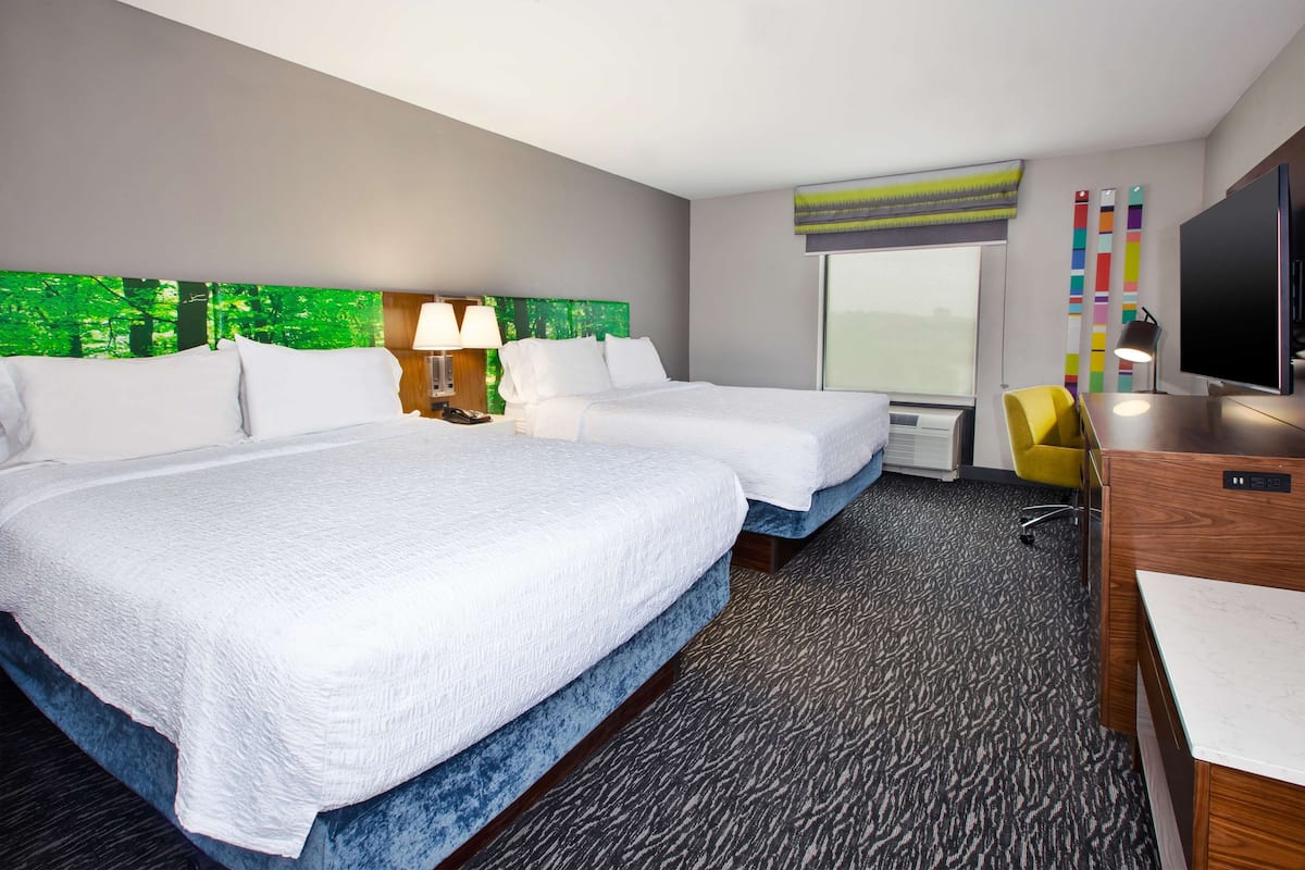 A hotel room with two beds, a desk, a TV, and a window with a view of trees.