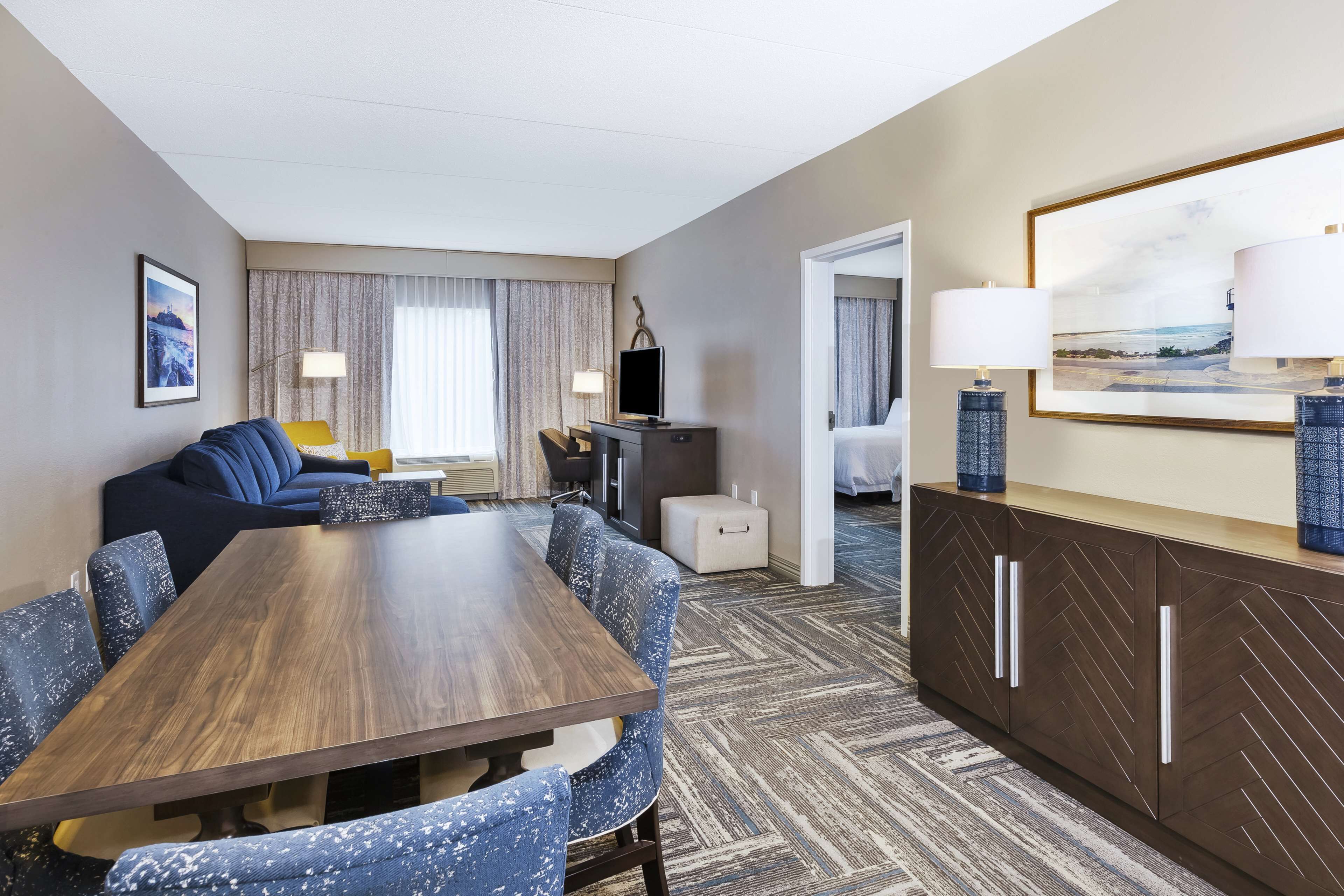 A hotel room with a wooden dining table, blue upholstered chairs, a sofa, a TV, a lamp, a bed, and a large framed picture on the wall.