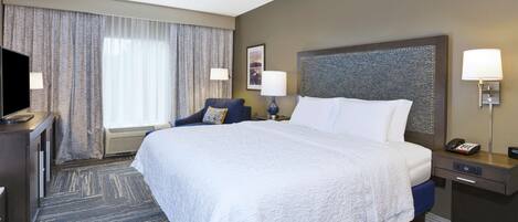 A hotel room with a large bed, a desk with a TV, a chair, and a nightstand with a lamp.