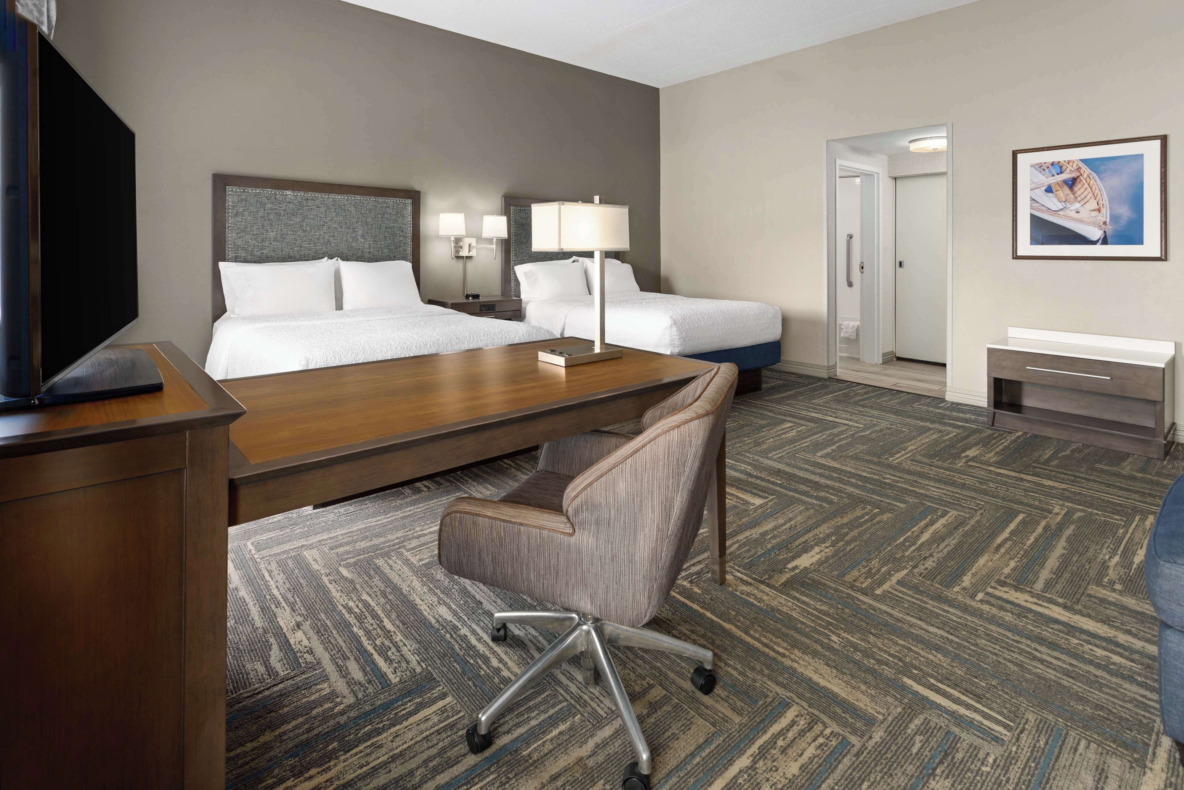 A hotel room with two beds, a desk, a chair, a TV, and a painting on the wall.