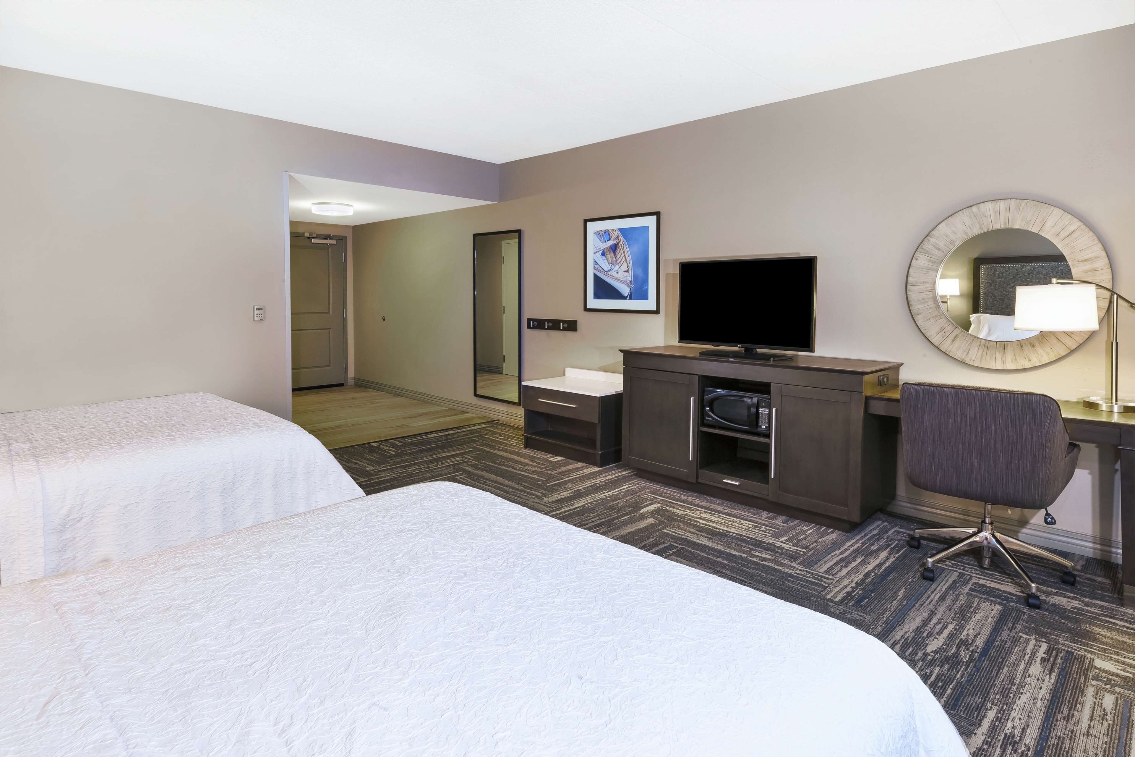 A hotel room with two beds, a desk with a chair, a television, a mirror, and a framed picture on the wall.