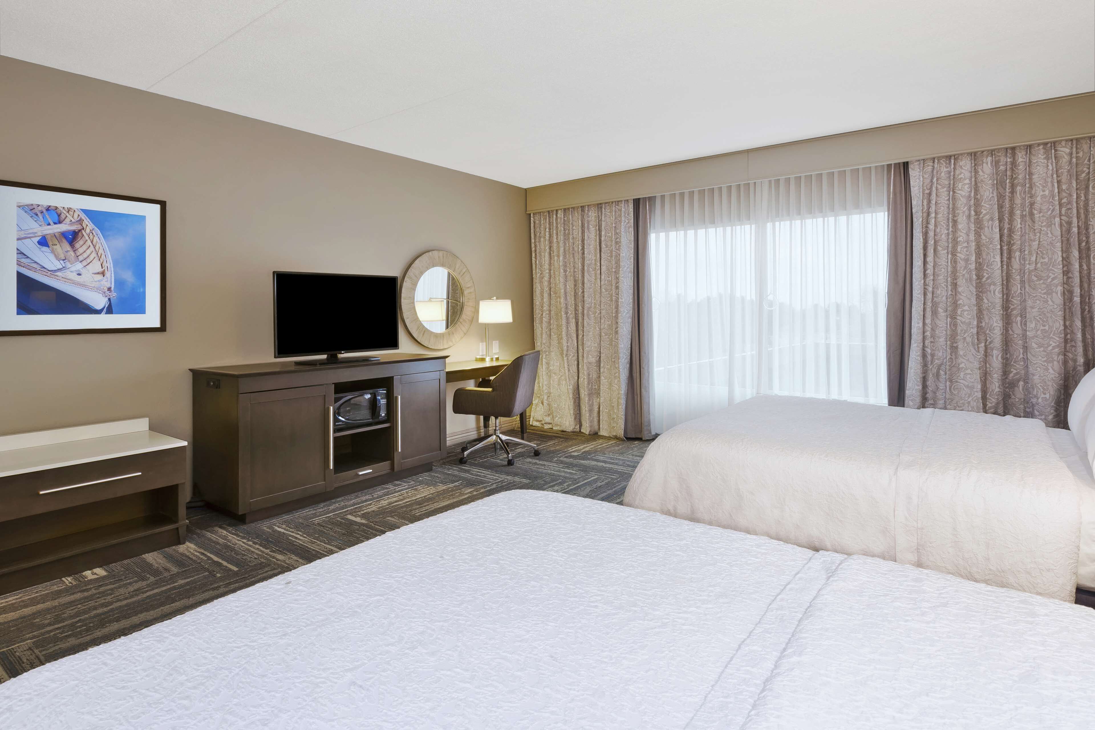 A hotel room with a bed, a desk with a chair, a TV, a mirror, and a window with curtains.