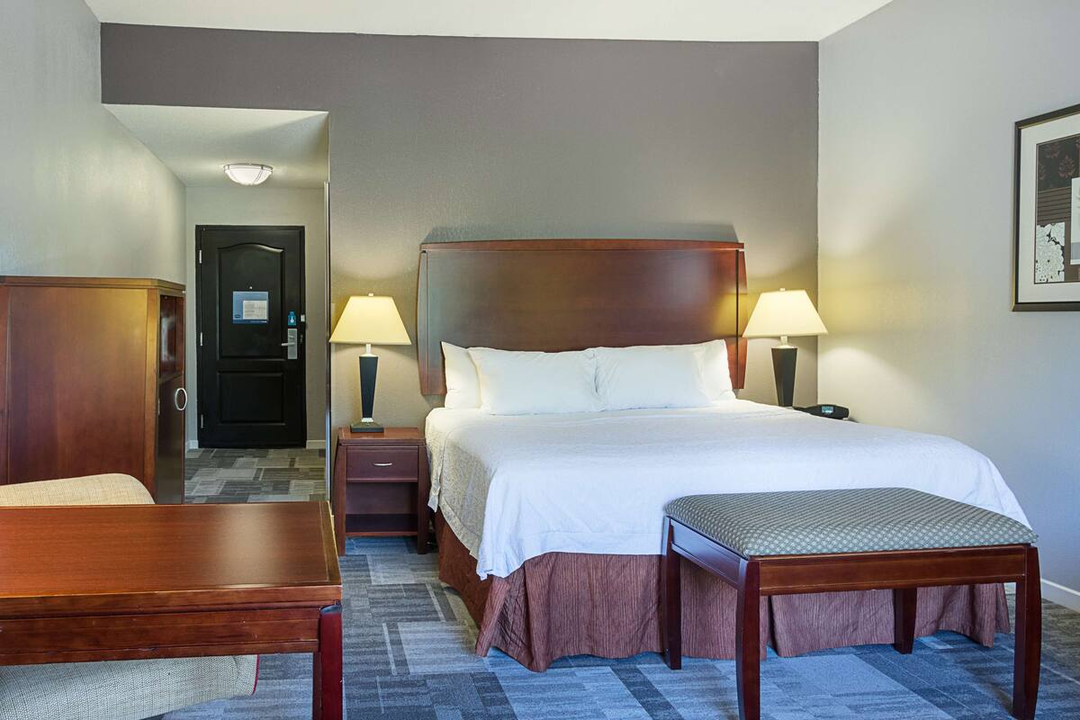 A hotel room with a large bed, two bedside tables with lamps, a wooden bench, and a door leading to a hallway.