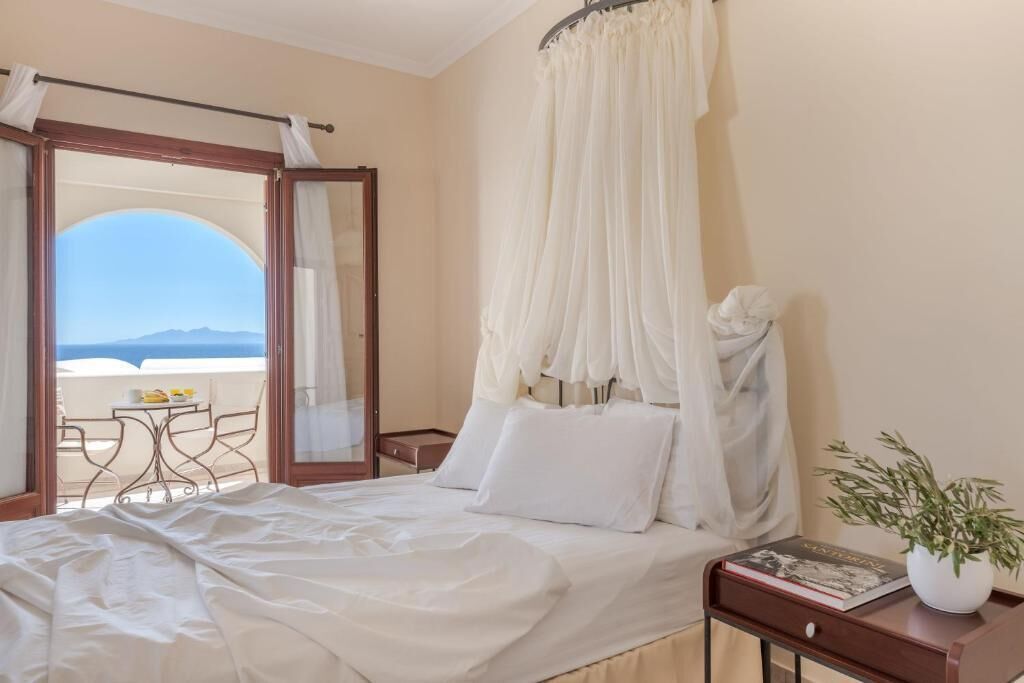 A hotel room with a bed, a bedside table with a book, a potted plant, and a view of the sea through an open door.