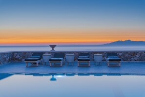 Zona de piscina con sillones de descanso y un muro de piedra, con vista a un mar tranquilo y montañas distantes al atardecer.