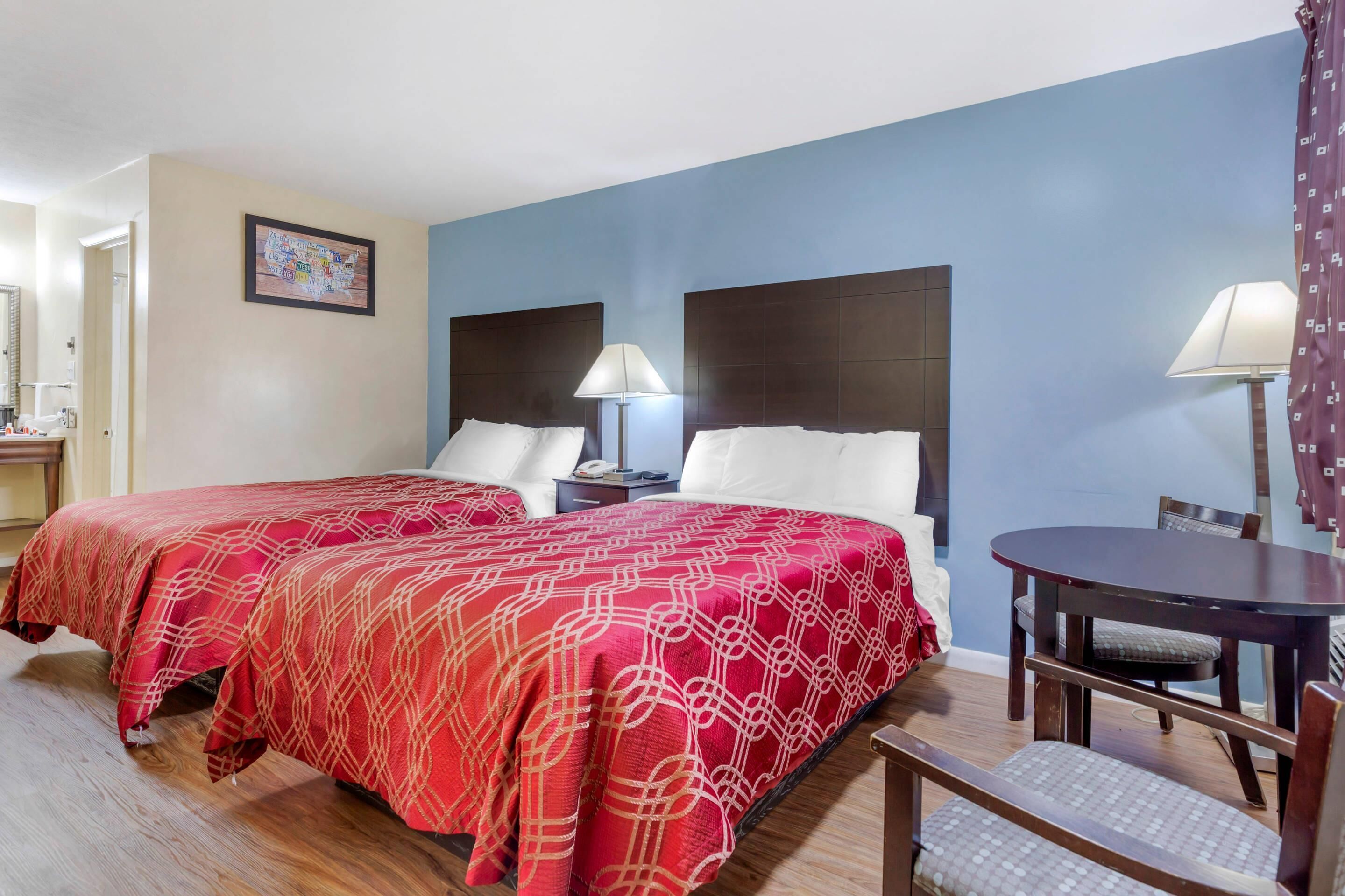 A hotel room with two beds, a dining table, a chair, a lamp, and a framed picture on the wall.