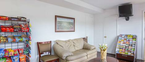 Una habitación con un sofá color beige, una silla de madera, una mesita redonda pequeña, un televisor montado en la pared, un estante para aperitivos y un tablón de anuncios.