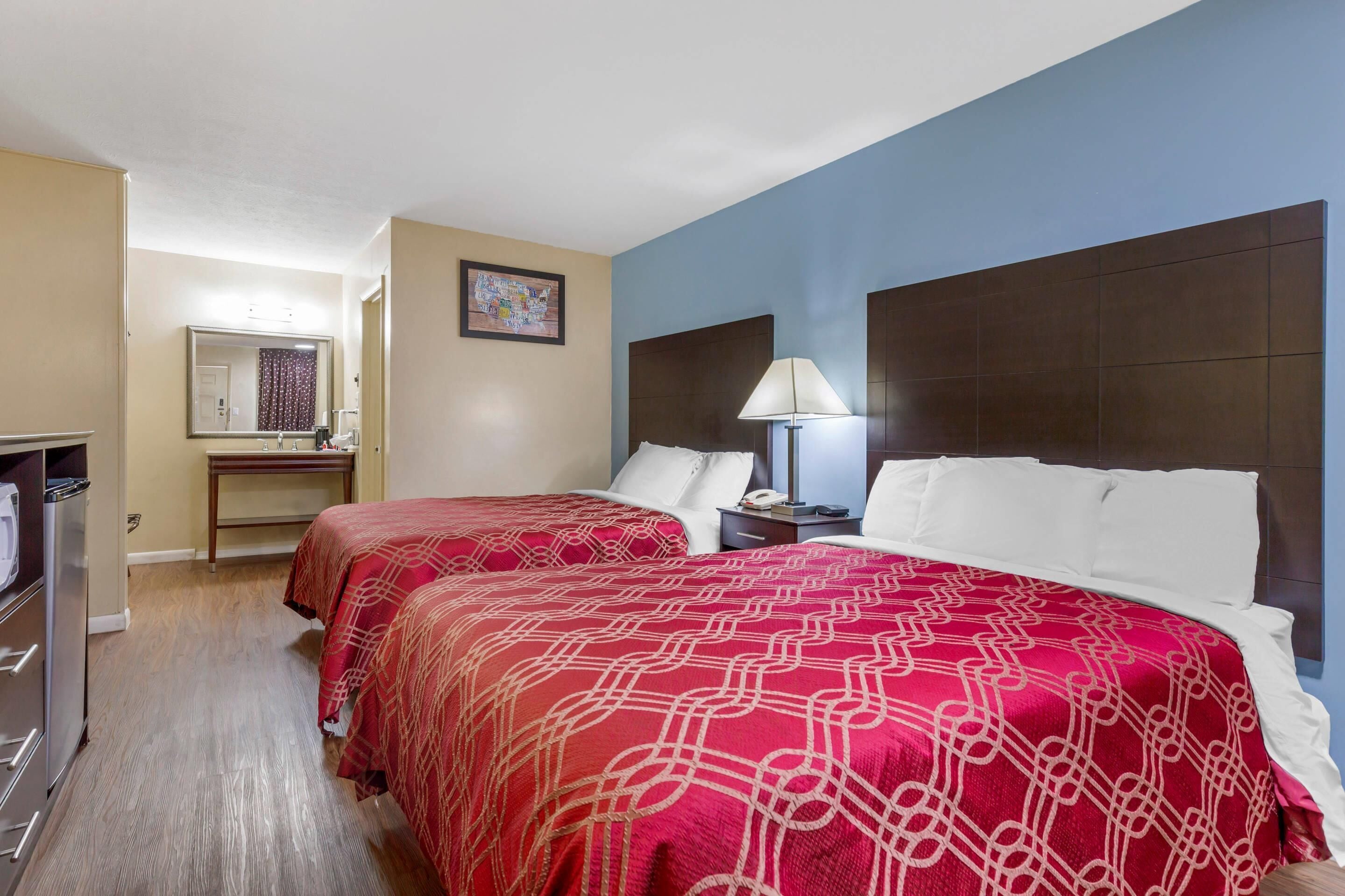 A hotel room with two beds, a desk, a mirror, and a nightstand with a lamp.