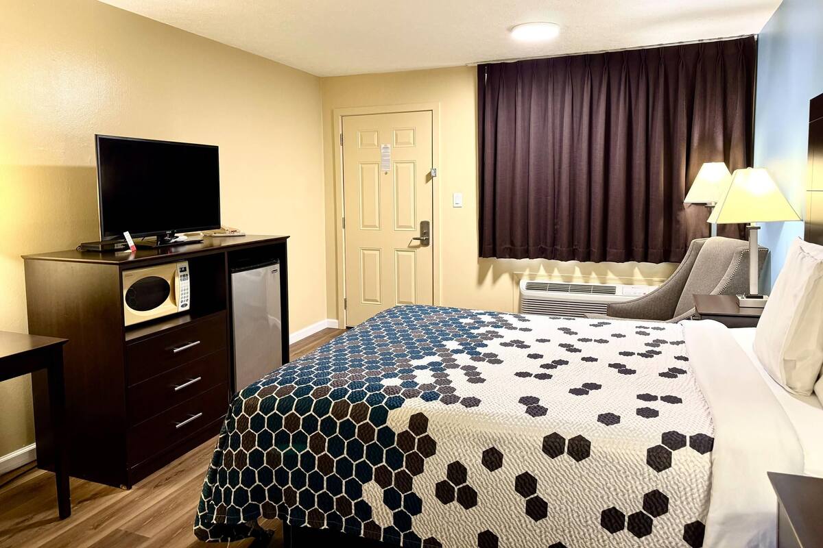 Ein Hotelzimmer mit einem Bett, einem Schreibtisch mit Fernseher, einem Sessel, einem Fenster mit Vorhängen und einer Lampe.