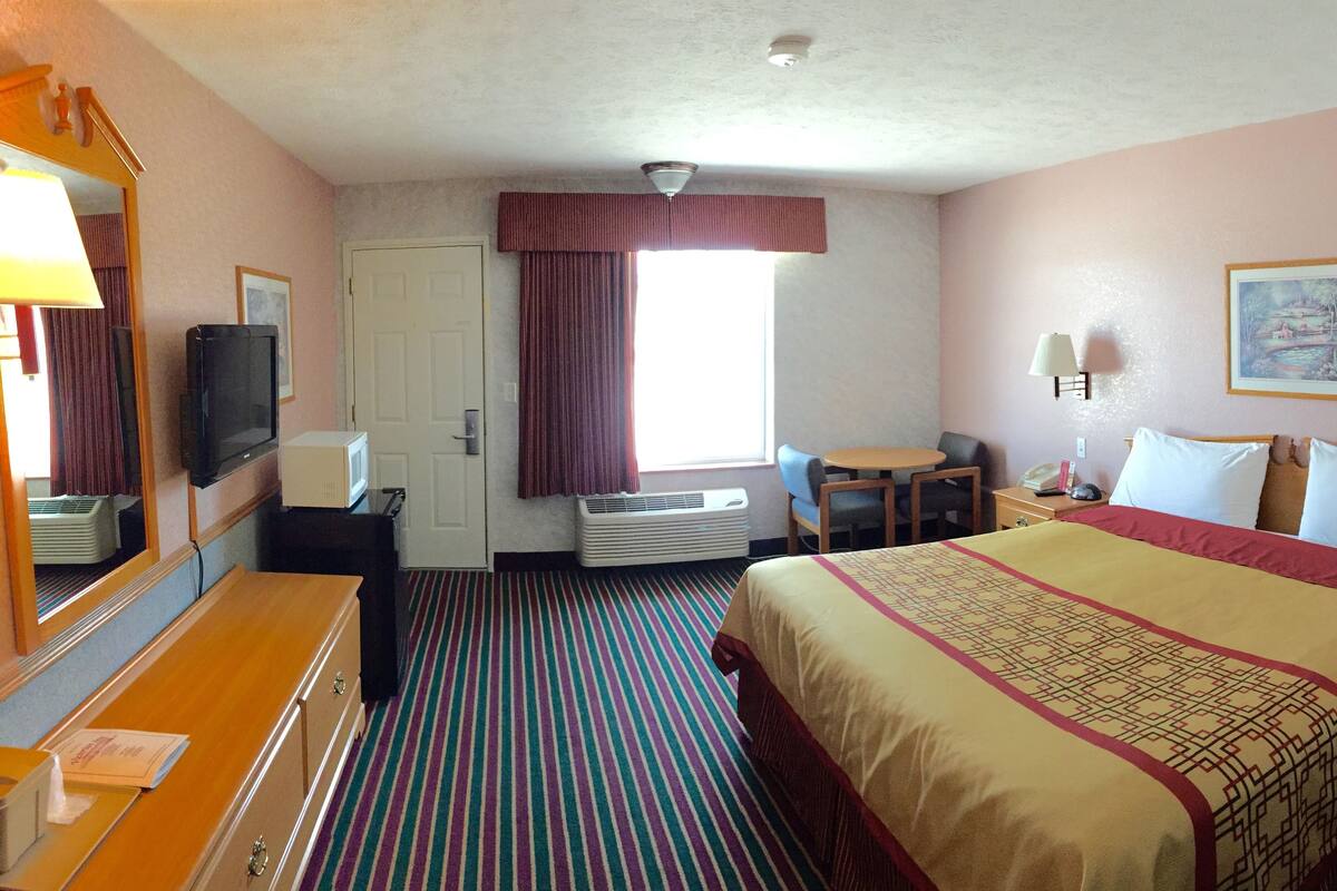 Quarto de hotel com cama, escrivaninha com cadeira, televisão e janela com cortinas.