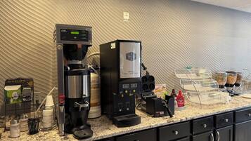 A coffee station with a coffee machine, a milk frother, and a coffee dispenser. There are condiment dispensers and a beverage cooler on the counter.