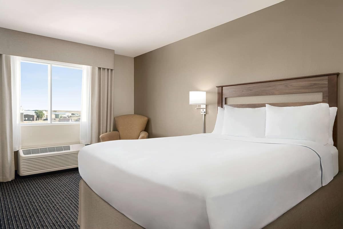 A hotel room with a large bed, a chair, a nightstand with a lamp, and a window with curtains.