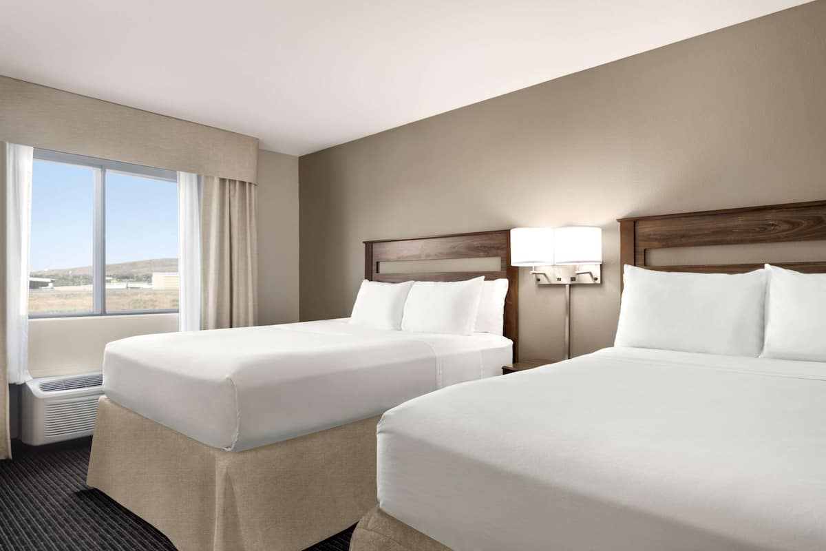A hotel room with two beds, a large window with curtains, a wall-mounted lamp, and a view of the outdoors.