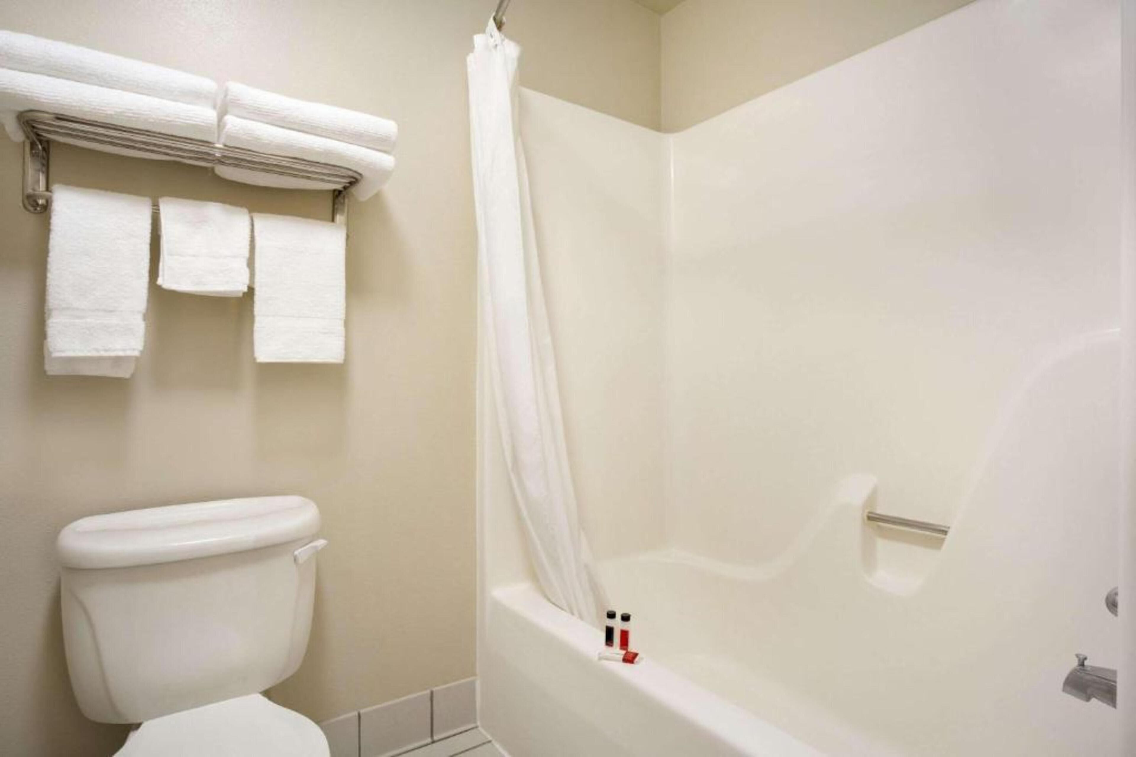 Een badkamer met een wit toilet, een douche met een witte gordijn en een handdoekrek met drie witte handdoeken.