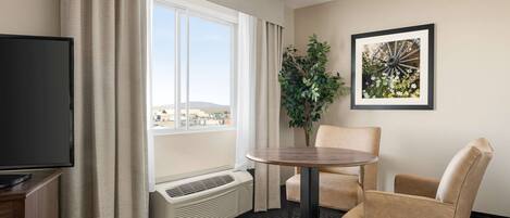 Ein Hotelzimmer mit großem Fenster, Flachbildfernseher, rundem Tisch, zwei beigen Sesseln, einem Topf mit Pflanze und einem gerahmten Bild an der Wand.