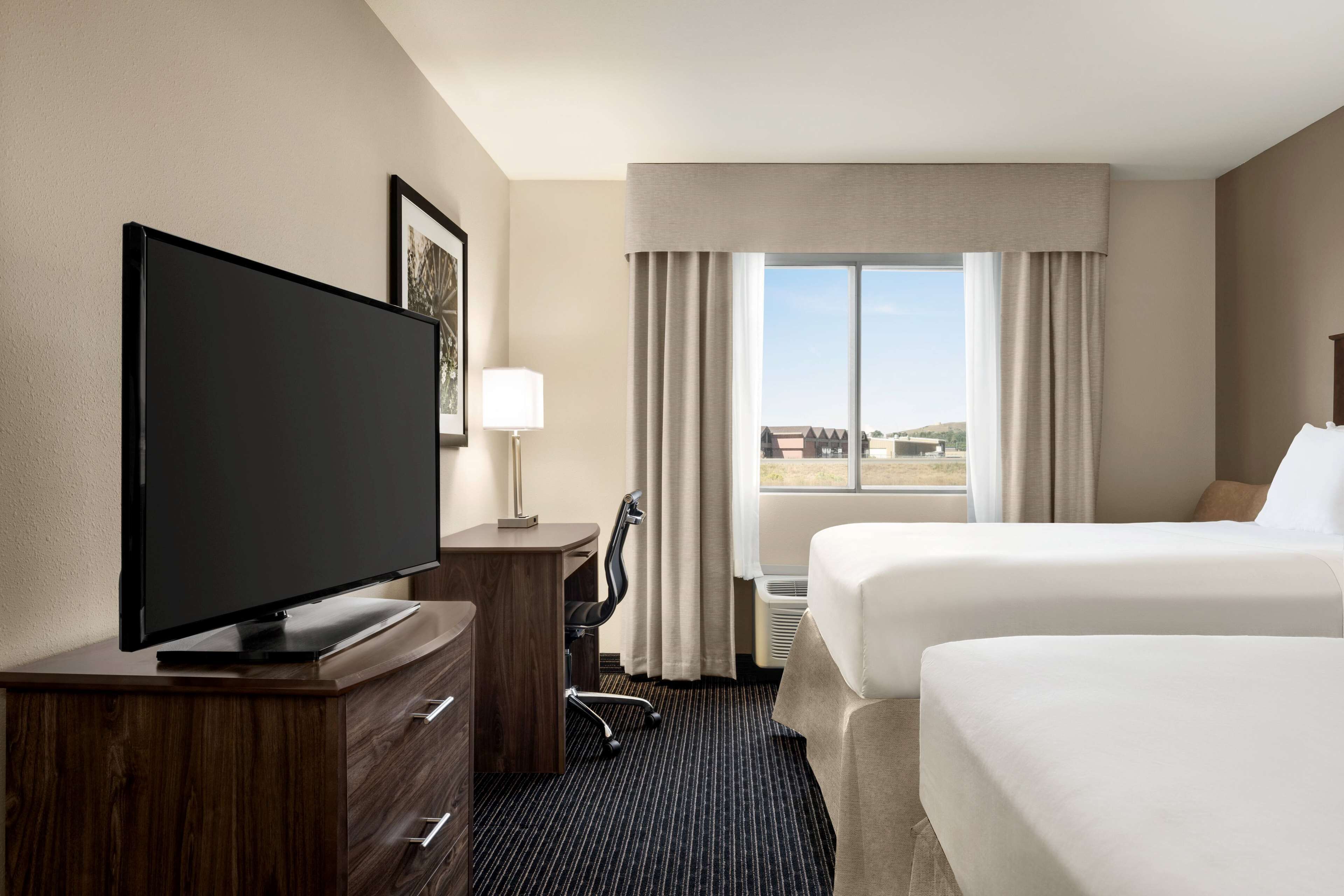 Ein Hotelzimmer mit Flachbildfernseher, Schreibtisch mit Lampe, zwei Betten und einem großen Fenster mit Vorhängen.