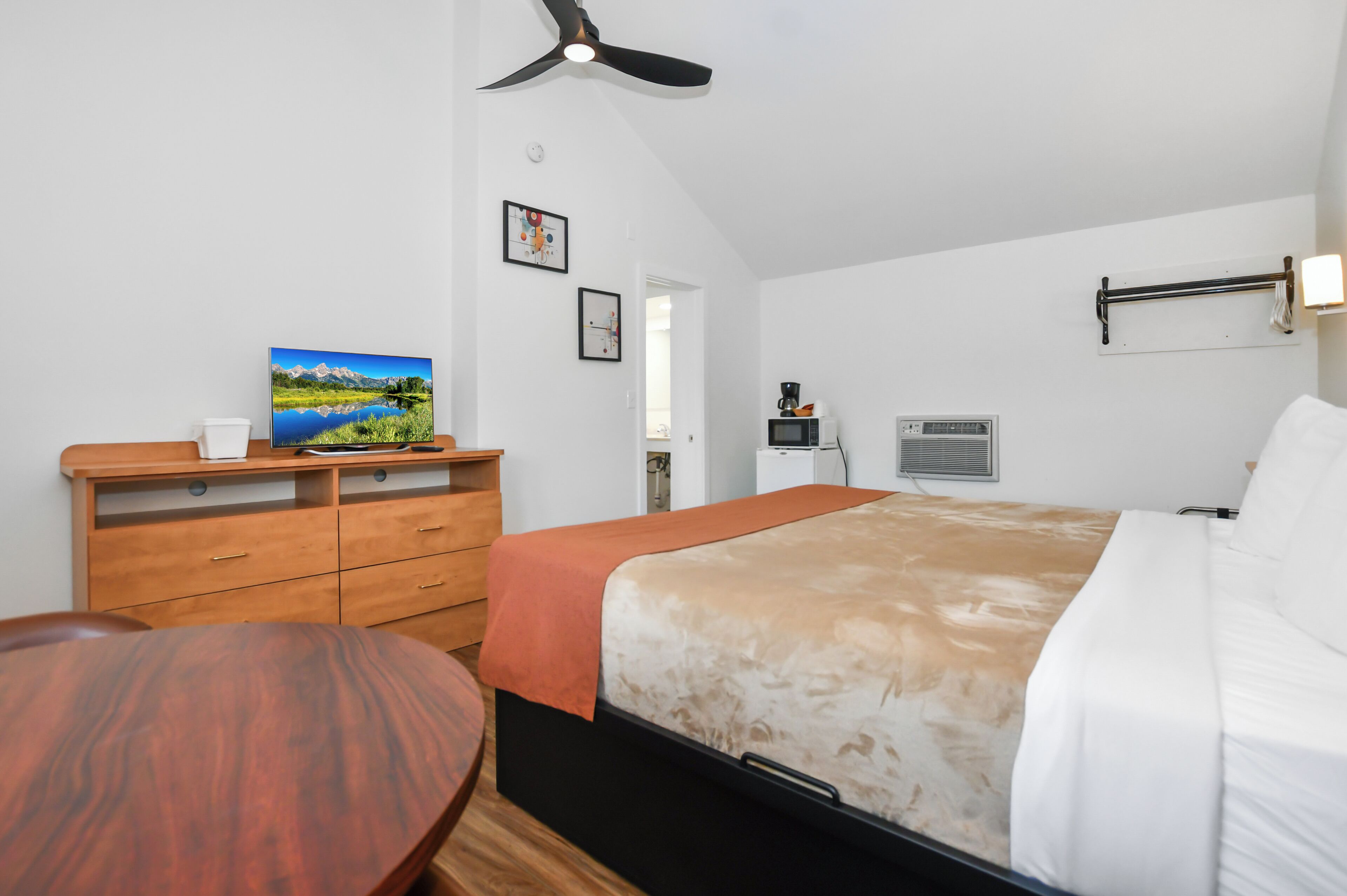 Un dormitorio con cama, cómoda de madera, ventilador de techo, televisor de pared y ventana.